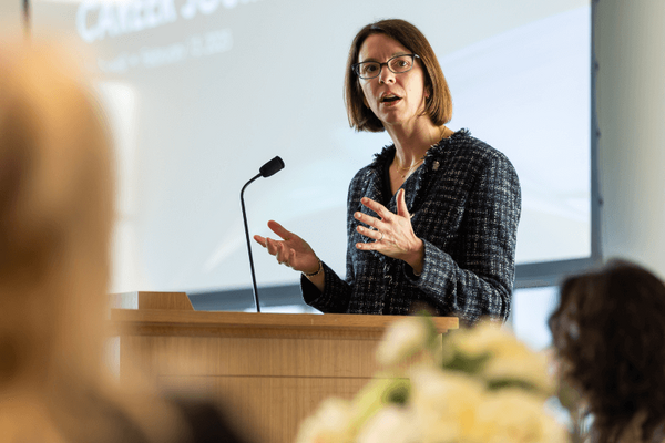 Kim Powell gestures with her hands as she speaks at a wooden podium. A microphone is positioned in front of her. Behind her, a projected screen displays the words "Career Journey."