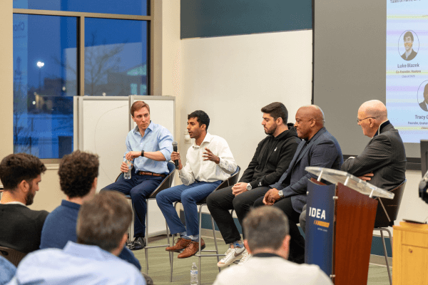 Five men sit on a panel facing the audience, engaged in a discussion.  A man in the center holds a microphone and gestures while speaking. An audience of students watches and listens.  A lectern with the IDEA Center logo is partially visible.