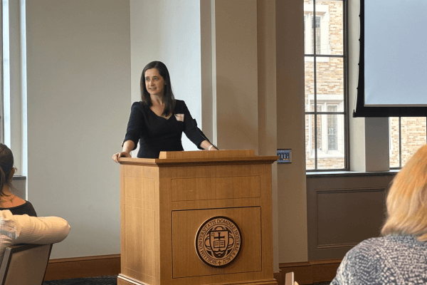 A woman in a black dress speaks at a wooden podium bearing the University of Notre Dame seal.  Audience members are partially visible.