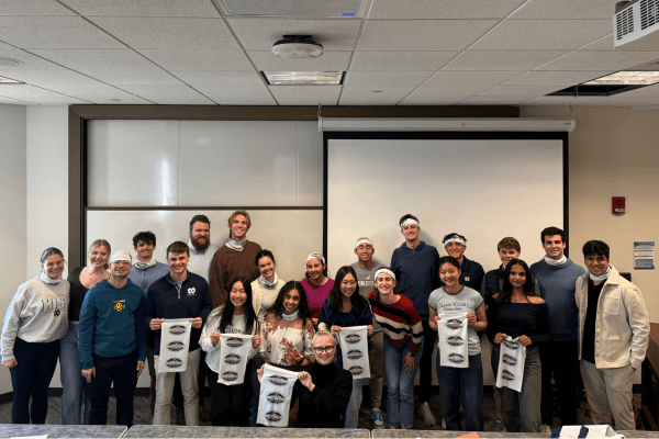 A group of students poses for a photo in a classroom, many holding small, white buffs with a blue and gold Survivor ND edition logo.  Several students wear them as headbands.  A whiteboard and projection screen are visible in the background.