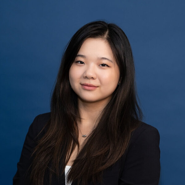 Olivia Cai headshot. A young woman with long dark hair, wearing a black blazer and a white top, smiles gently against a solid blue background.