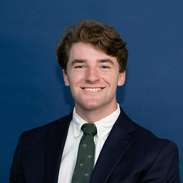 William Hamilton headshot. Young man with wavy brown hair and blue eyes, smiling broadly. He wears a navy suit jacket, white shirt, and dark green tie with a subtle horse pattern.