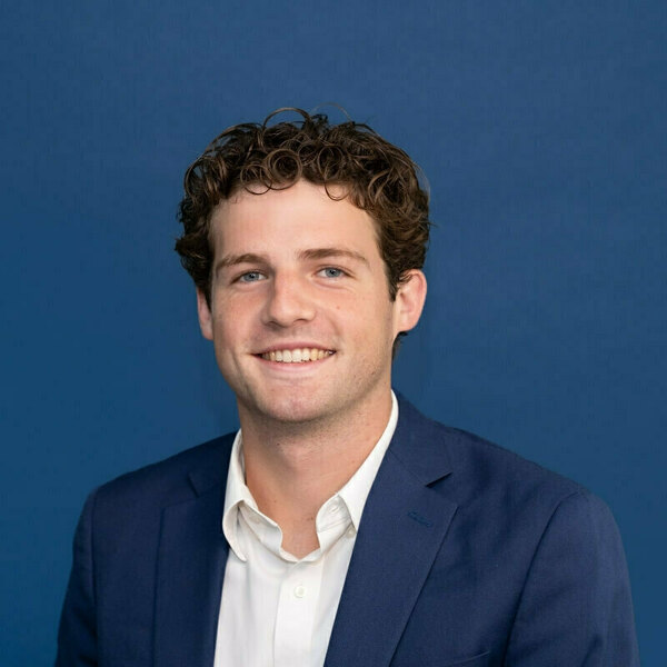 Will Favorite headshot. Young man with curly brown hair and blue eyes smiles against a blue background. He wears a white collared shirt and a navy blue blazer.