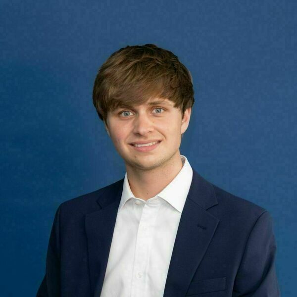 Ted Cahill Headshot A young man with brown hair and blue eyes smiles, wearing a white shirt and a dark blue blazer against a plain blue background.