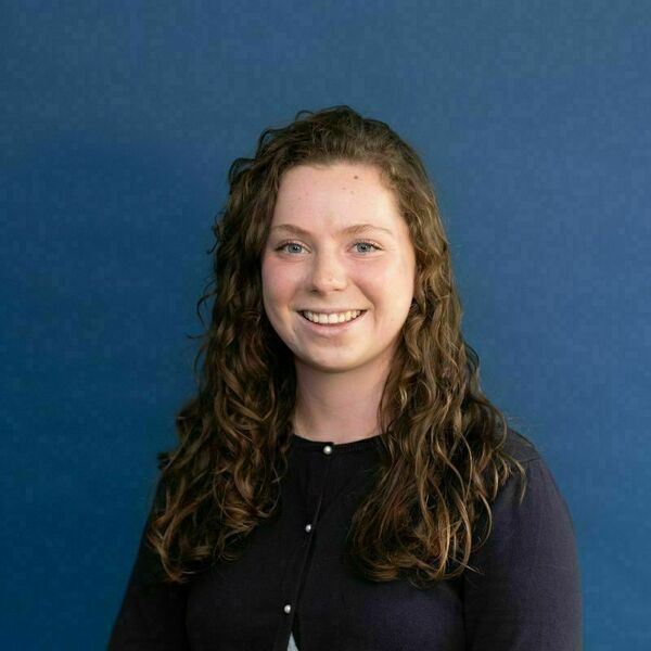 Violet McCann Headshot A young woman with long, curly auburn hair and blue eyes smiles broadly, showing her teeth. She wears a navy blue cardigan with light buttons, against a solid blue background.