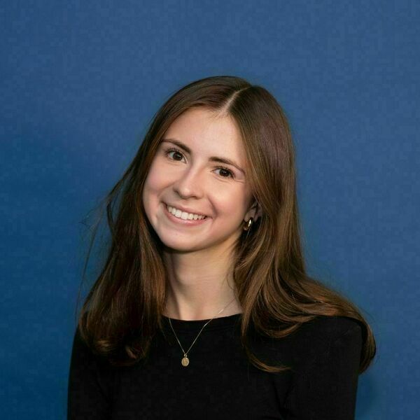 Anna O'Brien Headshot A young woman with long brown hair smiles broadly, wearing a black shirt, gold hoop earrings, and a gold pendant necklace against a blue background.