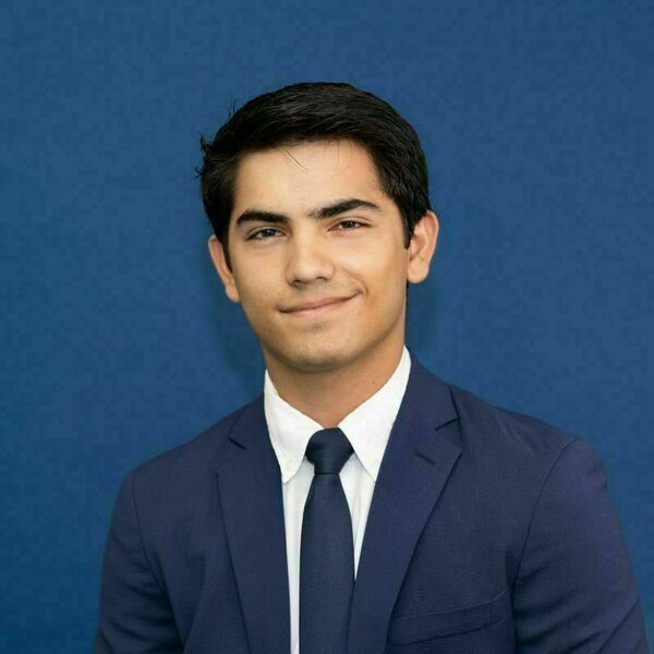 Bruce Alvarez Headshot A young man with dark hair and a slight smile wears a navy suit, white shirt, and navy tie against a blue background.