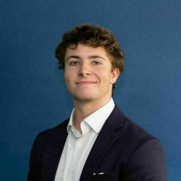Mac Sims Headshot. Young man with light brown curly hair and blue eyes, smiling slightly while wearing a navy blazer and white shirt against a blue background.