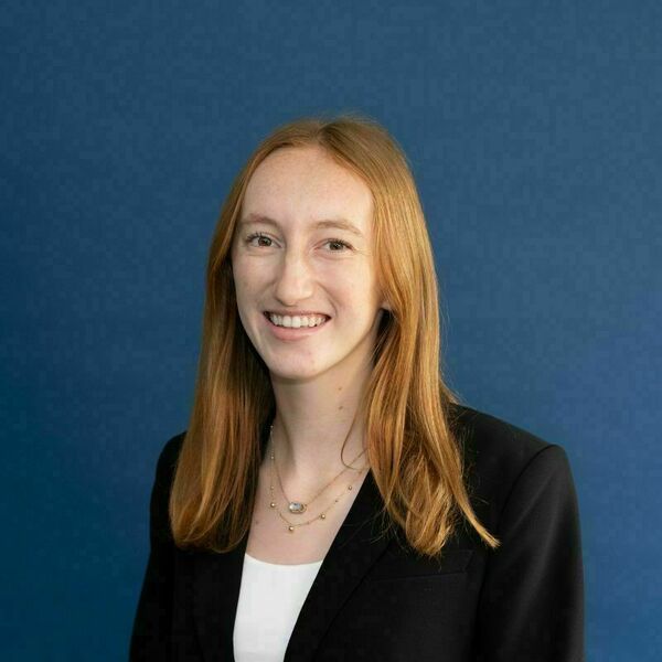 Julia Flood Headshot. A young woman with long reddish-blonde hair and freckles smiles, wearing a black blazer over a white top and layered gold necklaces against a dark blue background.