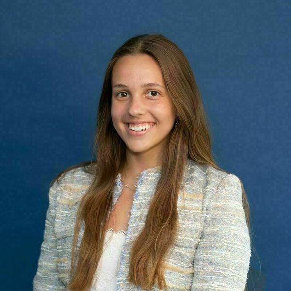 Maya Parafiniuk Headshot. Young woman with long brown hair smiles, wearing a textured light blue and beige jacket over a white lace top, against a solid blue background.
