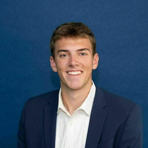 Charlie Thomas Headshot Young man with light brown hair and blue eyes, smiling broadly in a navy blue blazer and white shirt against a dark blue background.