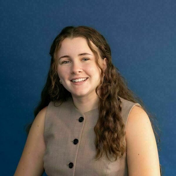 Molly Sullivan Headshot. Young woman with long wavy brown hair and gold hoop earrings, smiling broadly in a beige sleeveless top with dark buttons against a blue background.