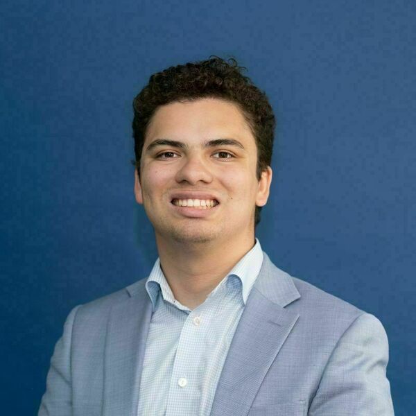 Nathan Ward Headshot. Young man with dark curly hair smiles, wearing a light blue checkered shirt and blazer against a deep blue background.