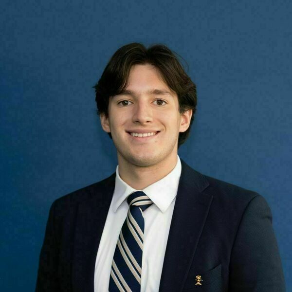 Paul Kaminski Headshot. Smiling young man with brown hair wears a navy suit, white shirt, and navy/beige striped tie, with a gold lapel pin, against a blue background.