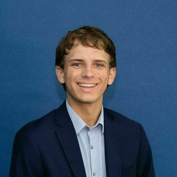 Raymond Warth Headshot. A young man with brown curly hair, blue eyes, and a broad smile, wearing a navy blue blazer and light blue collared shirt against a solid blue background.