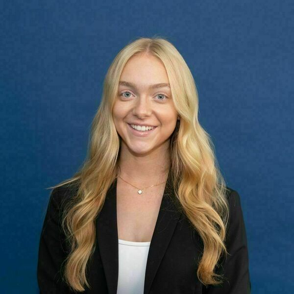 Sage Reeves Headshot. A young woman with long wavy blonde hair and blue eyes smiles brightly, wearing a black blazer, white top, and a delicate gold clover necklace, against a blue background.