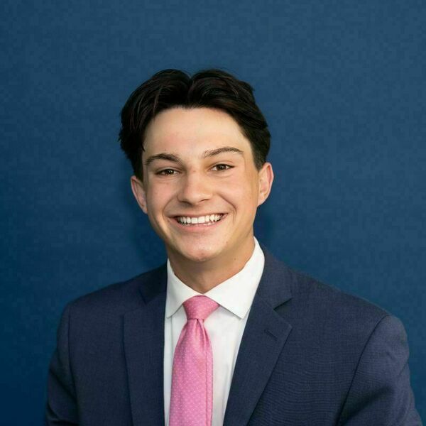 Daniel Mathy Headshot Young man with dark hair, wearing a dark blue suit, white shirt, and pink tie with white dots, smiles against a blue background.