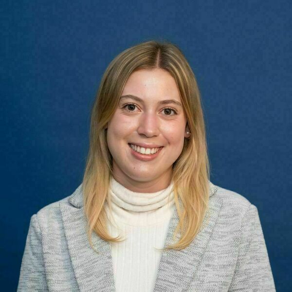Erin May Headshot Smiling young woman with long blonde hair, wearing a ribbed white turtleneck and a light gray blazer, in front of a blue background.