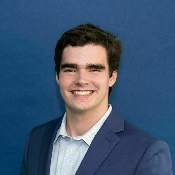 Jack Rankin Headshot Young man with dark hair and a wide smile, wearing a blue blazer and light blue shirt, against a solid blue background.