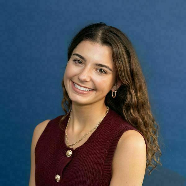 Adelyn Clemmer Headshot. A young woman with long brown wavy hair smiles broadly, wearing a burgundy button-up sleeveless top, a gold chain necklace, and gold hoop earrings against a blue background.