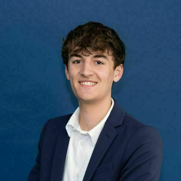 Drew Hemmesch Headshot. Young man with wavy brown hair, wearing a white shirt and navy blazer, smiling broadly against a blue background.