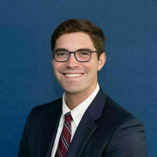Andrew Smith Headshot. Young man with brown hair and dark-rimmed glasses smiles against a dark blue background. He wears a dark blue suit, white shirt, and a maroon, navy, and white striped tie.