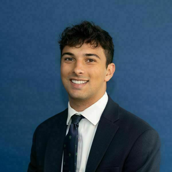 Ben Eppich Headshot. Smiling young man in a navy suit, white shirt, and a navy tie with green shamrock symbols, against a blue background.