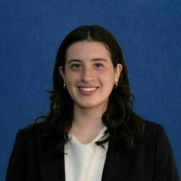 Catherine Paris Headshot. A young woman with dark wavy hair and freckles smiles broadly, wearing a white top and black blazer, against a royal blue background.
