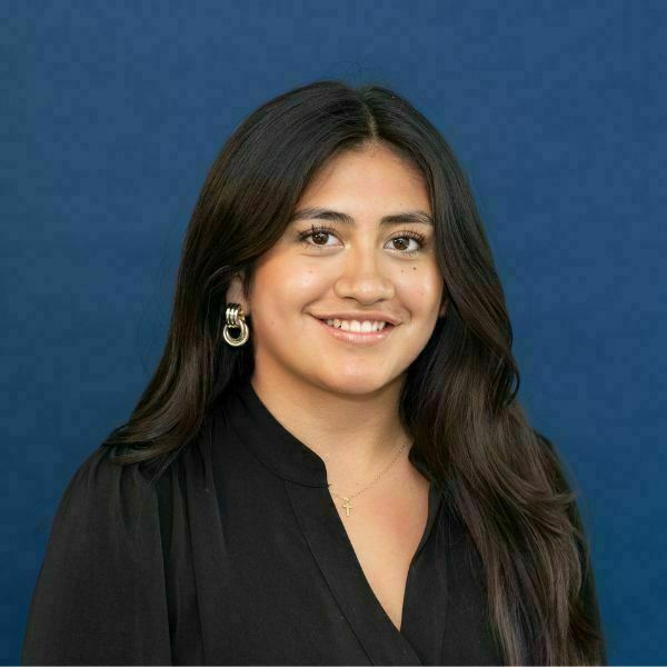 Daniella Morales-Garibay Headshot. A smiling young woman with long, dark, wavy hair, wearing a black blouse, gold double-hoop earrings, and a delicate gold cross necklace, against a royal blue background.