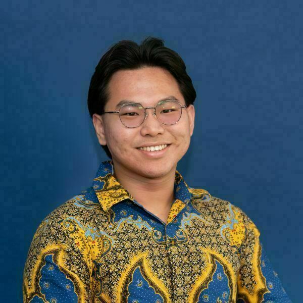 Darren Tanuwijaya Headshot. A young man with dark hair and round glasses smiles broadly, showing teeth. He wears a vibrant yellow and blue patterned shirt against a solid blue background.