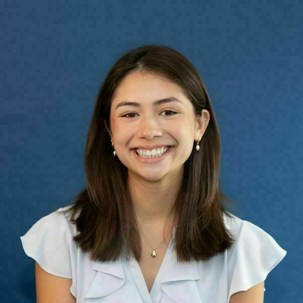 Grace Kloster Headshot. A young woman with long dark hair, wearing a light blue ruffled top, gold teardrop necklace, and pearl earrings, smiles broadly against a blue background.