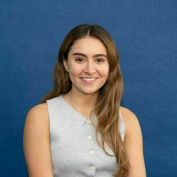 Isabella Uribe Headshot. A young woman with brown hair smiles, wearing a grey ribbed sleeveless top, against a blue background.