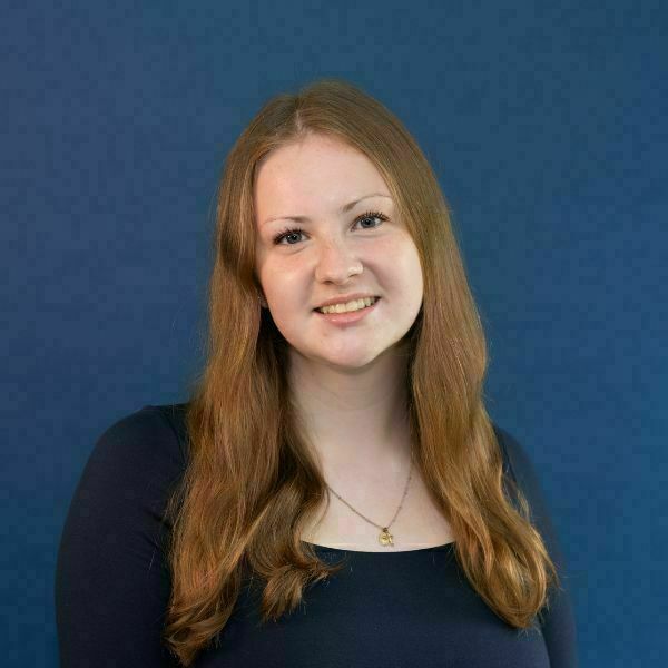 Katie Murphy Headshot. A young woman with long, reddish-brown hair, blue eyes, and a light complexion smiles against a dark blue background. She wears a dark blue shirt and a thin gold necklace.