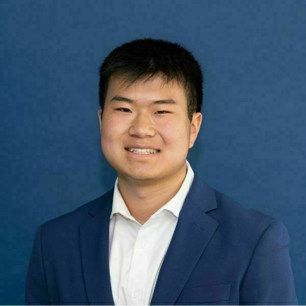 Kellan Ren Headshot. Smiling young man with short dark hair looks directly at the camera, wearing a navy blue suit jacket and a white collared shirt against a blue background.