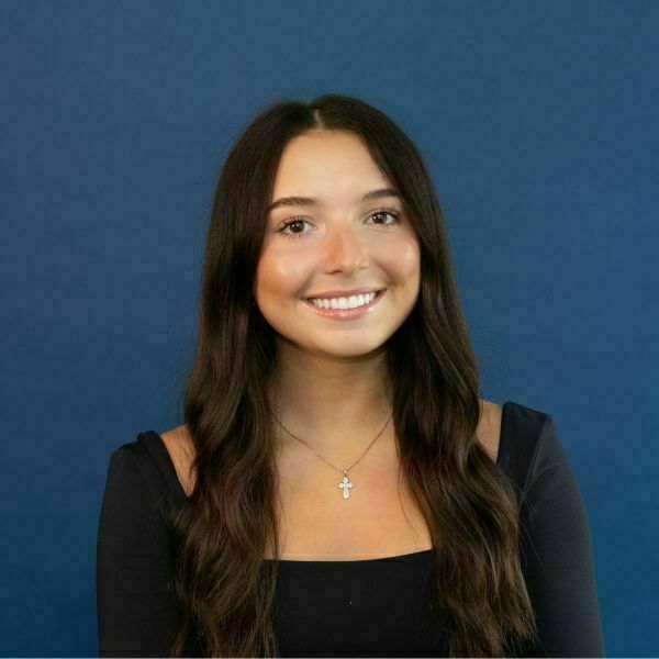 Kiki Petrovic Headshot. A young woman with long dark brown hair and brown eyes smiles broadly, wearing a black top and a silver cross necklace, against a blue background.