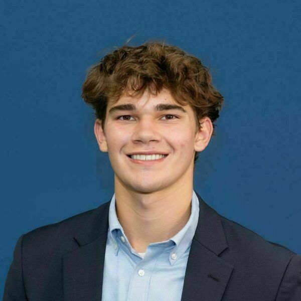 Joe Rozembajgier headshot. Young man with curly brown hair and a wide smile, wearing a navy blazer and light blue collared shirt against a solid blue background.