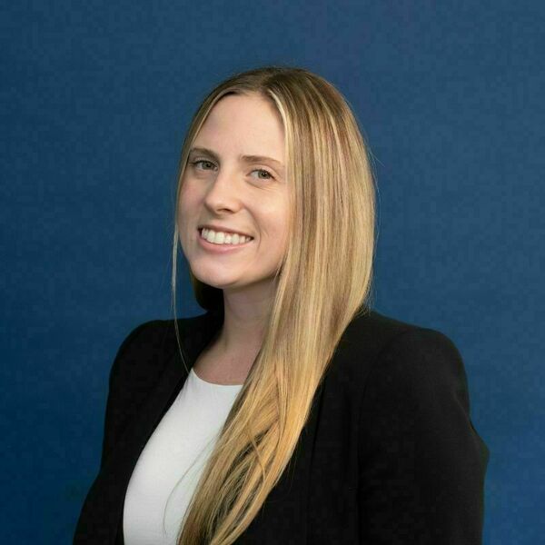 Julietta Tracy headshot. A young woman with long, straight blonde hair and a bright smile wears a black blazer over a white shirt, posing against a dark blue background.