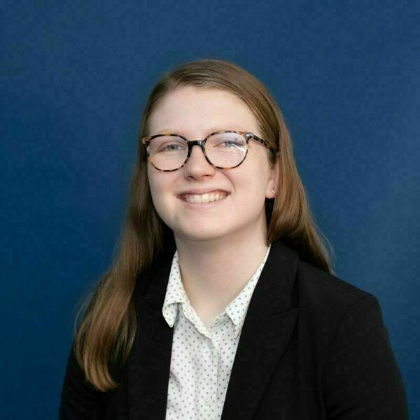 Lily Hagan headshot. Young woman with light brown hair and tortoise-shell glasses smiling against a dark blue background. She is wearing a patterned white shirt and black blazer.