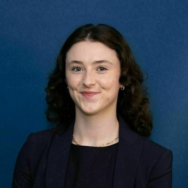 Margaret Abate headshot. A young woman with dark curly hair and blue eyes smiles gently, wearing a navy blazer and a gold necklace against a dark blue background.