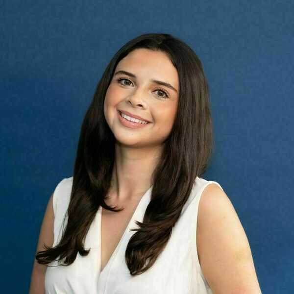 Alejandra Lopez headshot. Young woman with long, dark brown hair and a white sleeveless top smiles broadly against a solid blue background.