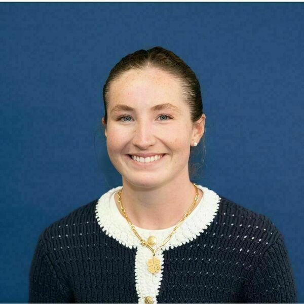 Maggie Minson headshot. Smiling young woman with freckles, light brown hair pulled back, and blue eyes, against a plain blue background. She wears a navy blue knit cardigan with white crochet trim and a gold necklace with a four-leaf clover pendant.