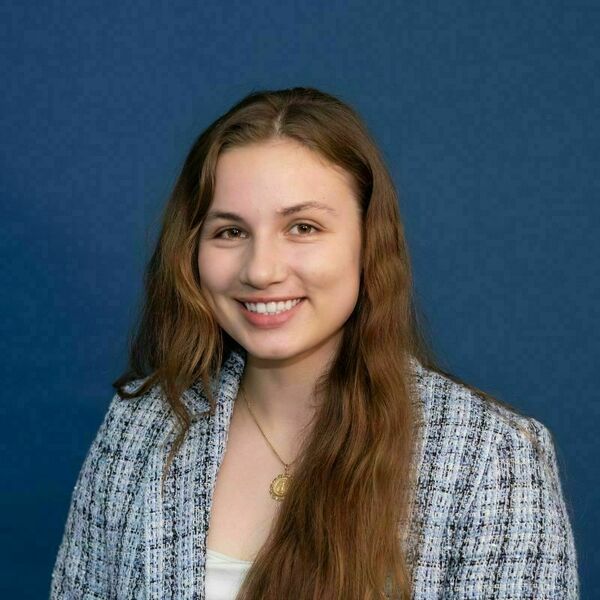 Leia Barth headshot. A young woman with long brown hair smiles broadly, wearing a light blue and white tweed jacket and a gold medallion necklace, against a dark blue background.
