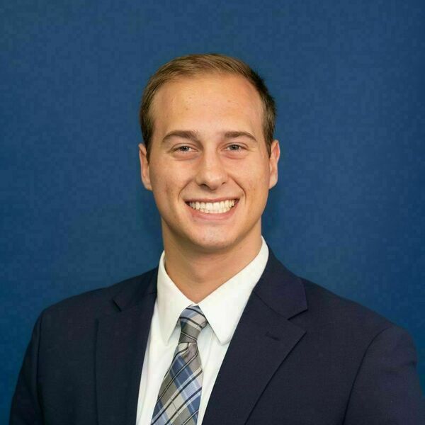 Jerry Vielhauer headshot. Young man with short brown hair, wearing a navy suit, white shirt, and blue plaid tie, smiling broadly against a dark blue background.