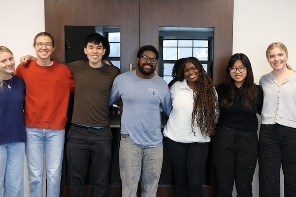 Seven 2026 student interns stand arm-in-arm against a white wall with dark doors. They wear various casual tops in blue, orange, brown, light blue, white, and black, with jeans or dark pants.