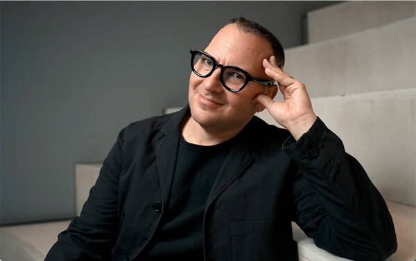 Man in black jacket and glasses smiles with hand near his head.